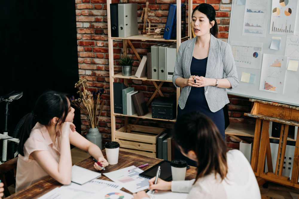 confident young pregnant female manager stand talk while addressing team board meeting office group women employees concentrated listening making note start up business concept