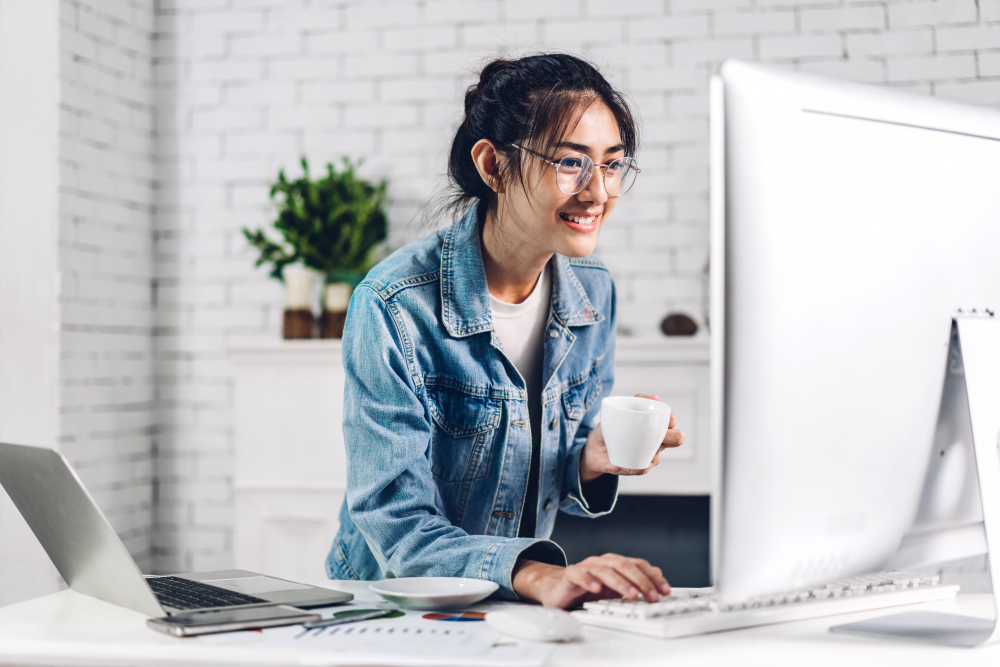 young smiling happy asian woman relaxing using desktop computer working video conference meeting online chat young creative girl drink coffee home work from home concept