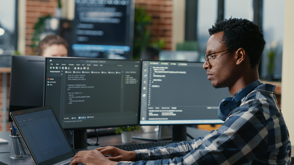 sofware developer thinking while touching beard while typing laptop sitting desk with multiple screens parsing code focused database admin working with team coding background